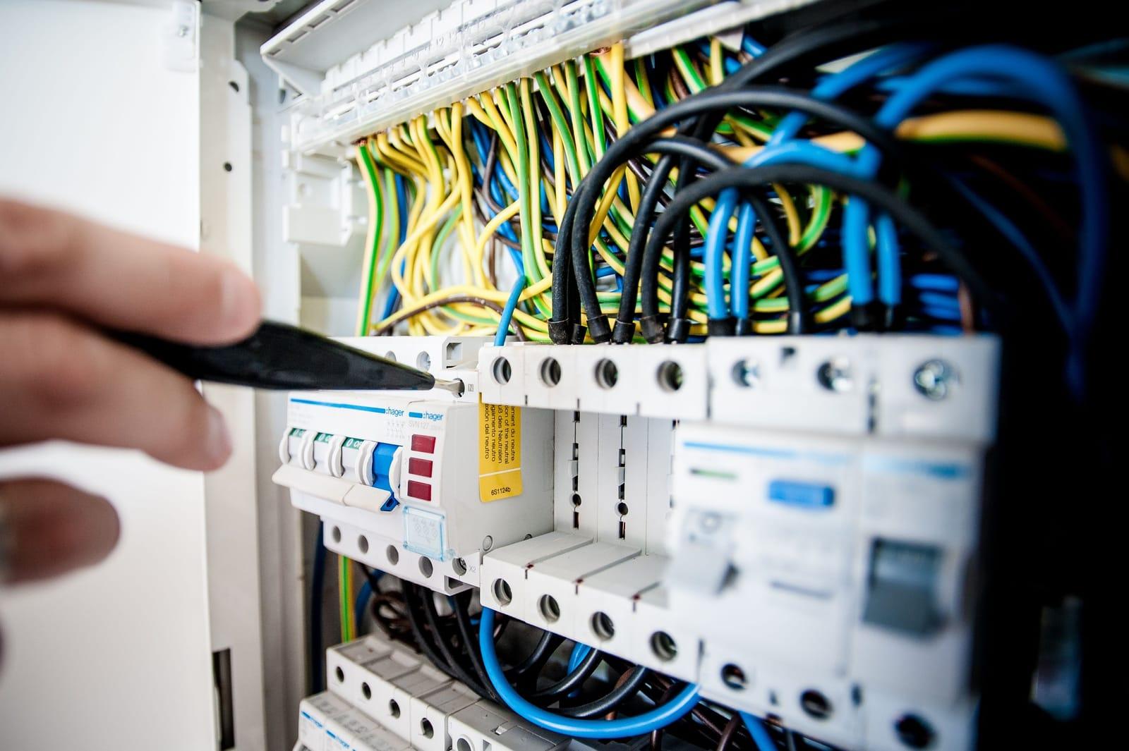 Mână cu șurubelniță care strânge o bornă într-un tablou electric deschis, cu siguranțe automate, diferențial și cabluri albastre, galben-verzi și negre trase deasupra